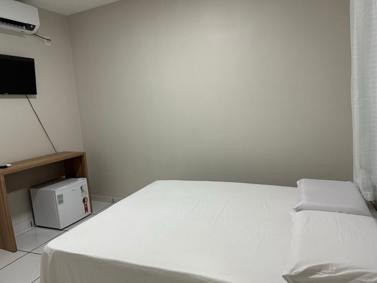 Quarto simples e limpo, com uma cama, uma pequena escrivaninha com frigorífico e uma televisão montada na parede.