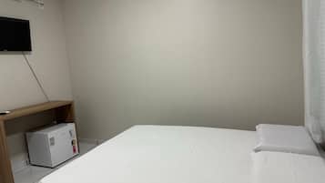 A simple, clean room with a bed, a small desk with a refrigerator, and a wall-mounted TV.