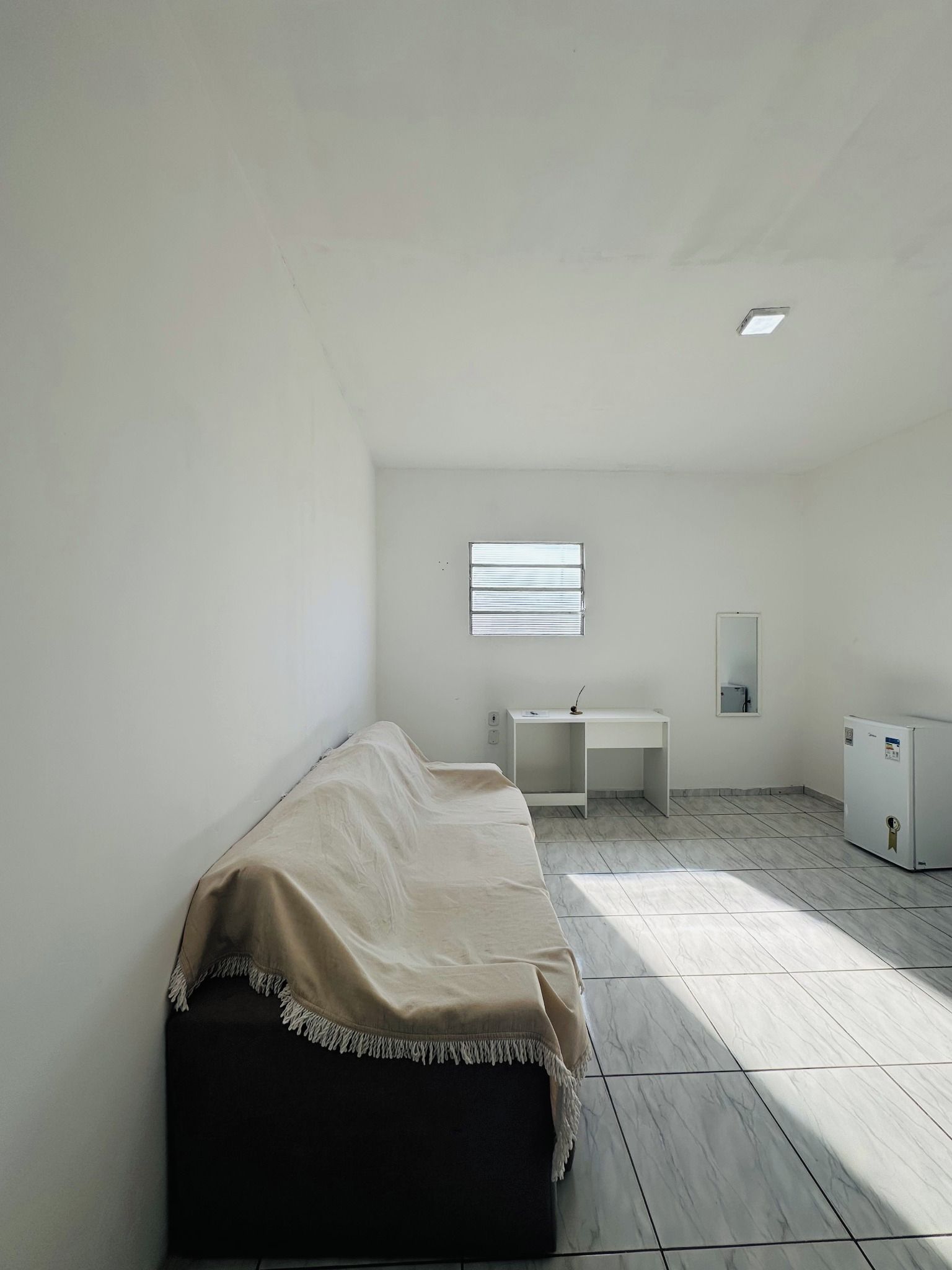 Una habitación pequeña y sencilla con cama, escritorio, espejo y refrigerador.