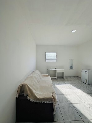 A small, unadorned room with a bed, a desk, a mirror, and a refrigerator. - Pousada Central dos Lençóis (Barreirinhas)