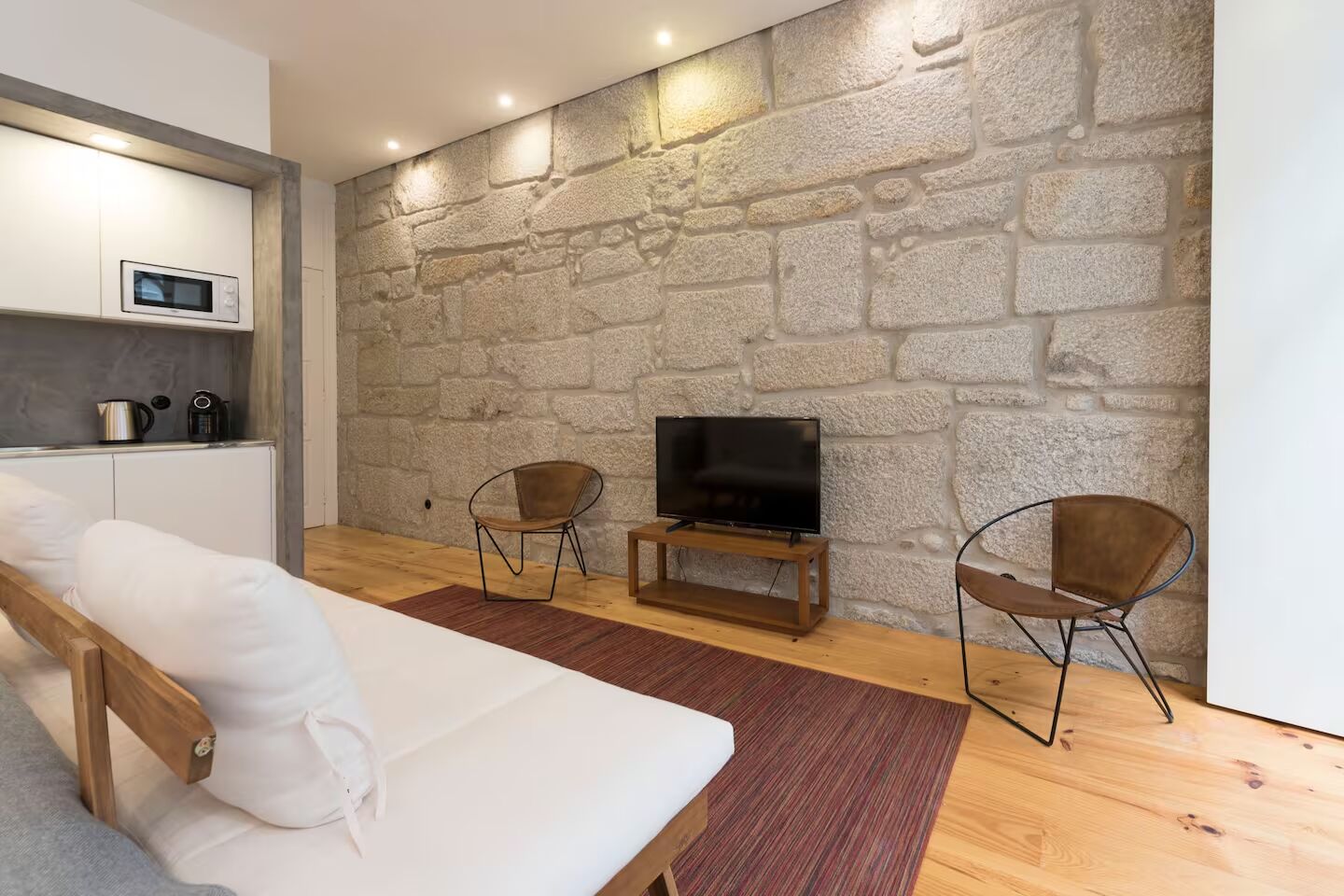A modern living room with a stone wall, a flat-screen TV, two chairs, a sofa, and a kitchen area with a microwave.