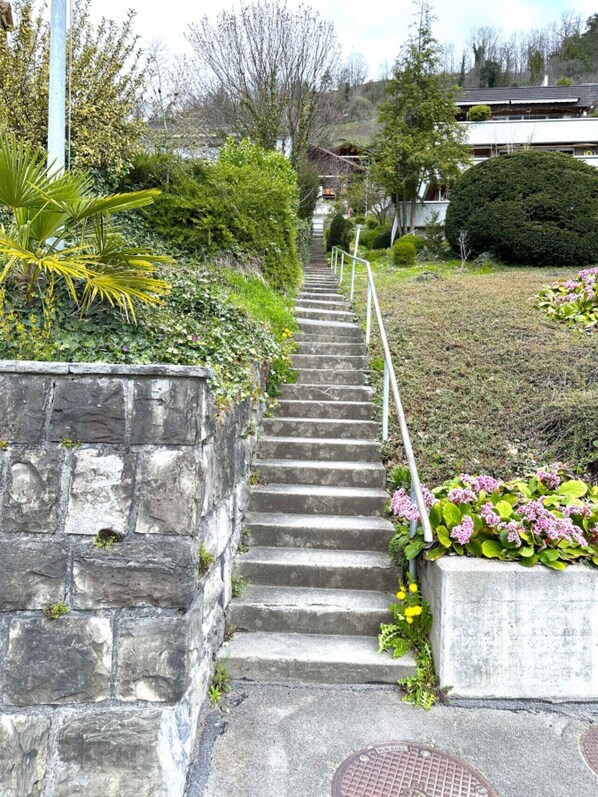 A set of stone steps leading up a hill, surrounded by greenery and a residential building in the background.