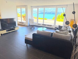 Ein modernes Wohnzimmer mit einer großen Couch, einem Flachbildfernseher und Seeblick durch große Fenster.