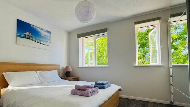 Un dormitorio con cama, dos ventanas, una mesita de noche y una lámpara de pared.