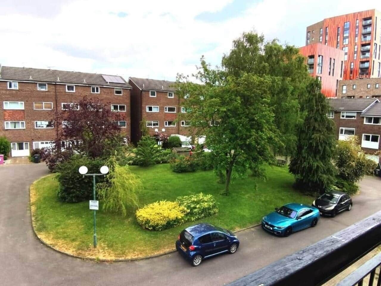 Ett bostadsområde med en rad tegelbyggnader, en grön gräsmatta med buskar och två bilar parkerade längs gatan.