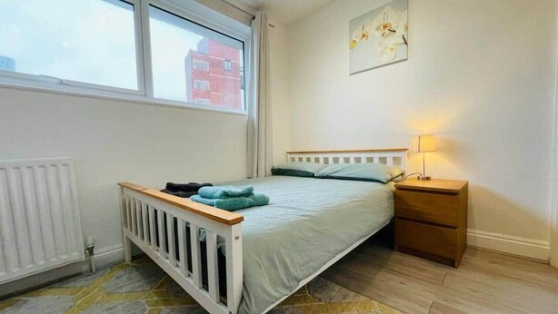 Een slaapkamer met een bed, nachtkastje, radiator, raam en een schilderij aan de muur.