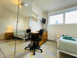 Ein Schlafzimmer mit Bett, Schreibtisch, Stuhl, Kommode, Spiegel, Fenster und Heizkörper.