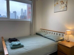 A single bed with a wooden headboard, a bedside table with a lamp, and a window with a cityscape view. - Nice bedroom with private bathroom in London Canary Wharf (London)