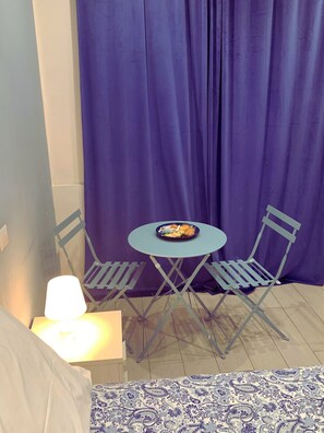 Una habitación pequeña con una cama, una mesa con sillas, una lámpara y una cortina.