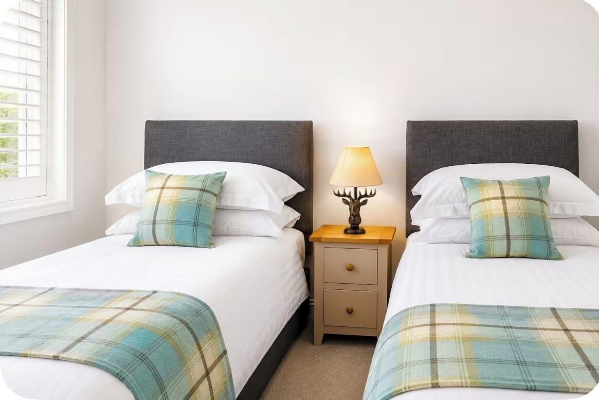 Een hotelkamer met twee bedden, elk voorzien van een ruitbeddeken, een bedlamp en een raam met luxaflex.