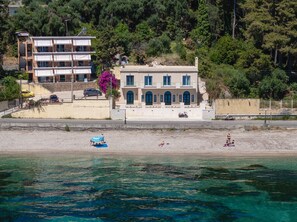 A coastal building with a balcony overlooking the sea, a sandy beach, and a forested hillside. - Luxury Beachfront Achillion Bay Villa | Infinity Pool & Breathtaking Sea Views (Mpenitses)