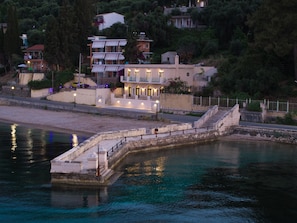A coastal hotel with a stone staircase leading to a beach, surrounded by lush greenery and other buildings. - Luxury Beachfront Villa with infinity Pool & Breathtaking Sea Views (Mpenitses)