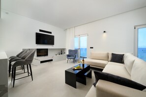 A modern living room with a flat-screen TV, a sofa, a coffee table, and a dining area with bar stools. - Luxury Beachfront Villa with infinity Pool & Breathtaking Sea Views (Mpenitses)