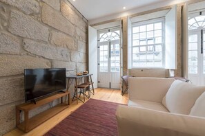 En stue med en stenvæg, et fladskærms-tv på et træstel, en hvid sofa, et lille bord med stole og store vinduer med udsigt til bygninger.
