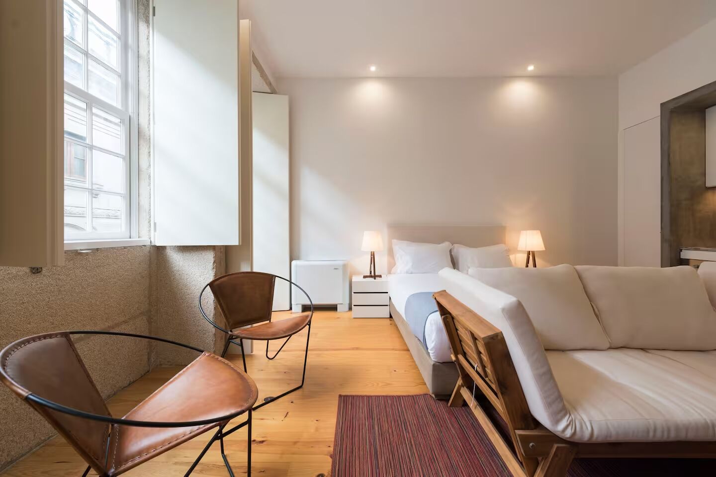 Una habitació d'hotel amb un llit, dues taules de nit amb làmpades, una cadira, un terra de fusta i una finestra.