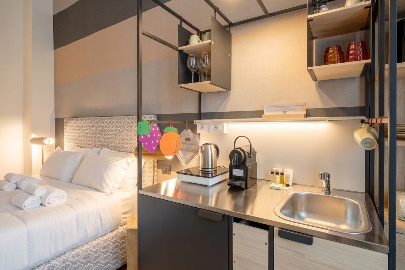 Habitación de hotel pequeña con cama, kitchenette, lavamanos y mesita de noche con tetera y hervidor de agua.