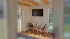 Quarto com teto de madeira, televisão de tela plana na parede, mesa redonda pequena com duas cadeiras e uma janela com persianas.