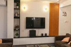 A modern living room with a flat-screen TV mounted on the wall, a wooden accent wall, and a built-in shelf with decorative items. - Luxury 2-Bedroom Apartment in Nyali with Pool & Beach Access in Mombasa (Mombasa)
