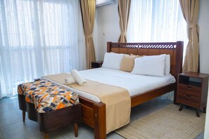 A hotel room with a wooden bed, a bedside table, a nightstand, and a large window with curtains. - Luxury 2-Bedroom Apartment in Nyali with Pool & Beach Access in Mombasa (Mombasa)