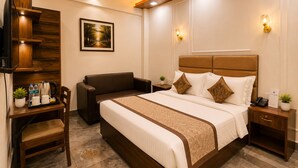 Ein Hotelzimmer mit einem Bett, einem Schreibtisch, einem Stuhl, einem Nachttisch, einem an der Wand montierten Fernseher und einem Deckenventilator.