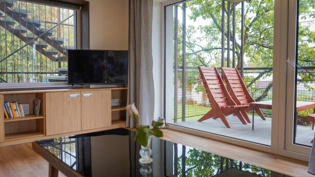 A room with a television on a wooden cabinet, a window with a view of outdoor furniture, and a vase with flowers on a windowsill.