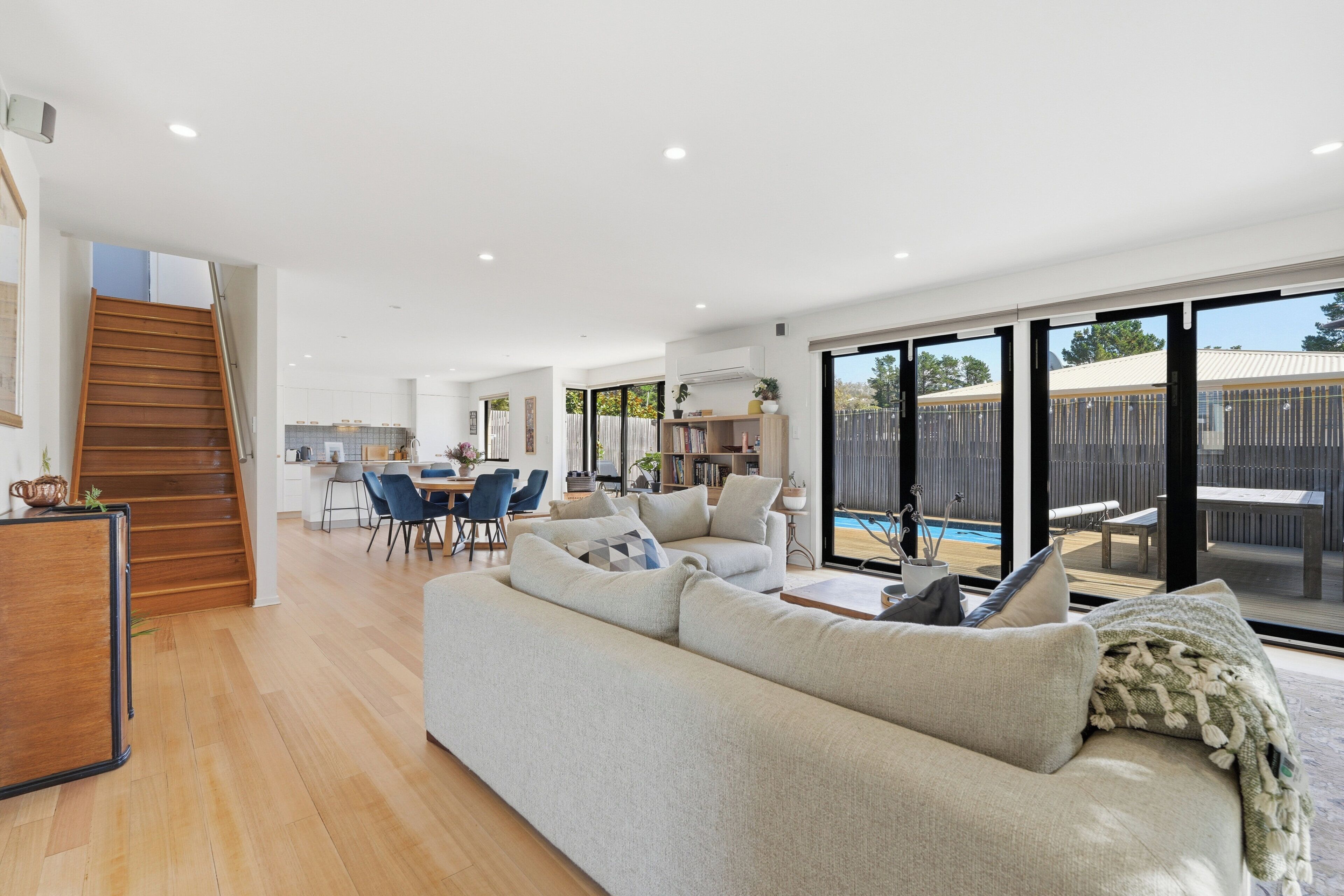 A spacious living area with a large sofa, dining table, and chairs, a staircase leading to an upper level, and a sliding glass door to a pool area.