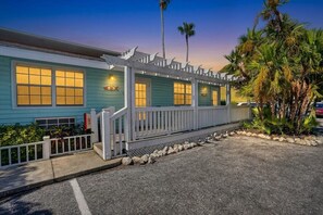 A building with a porch and a covered walkway, surrounded by palm trees and a gravel driveway. - Tradewinds #31 –  Walk to the Beach • Bayfront Community Pool (Bradenton Beach)