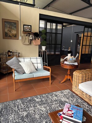 A living room with a sofa, a wicker chair, a small round table, and a bookshelf. - 8 @ Mill Lane • Hartbeespoort Guesthouse • Hot Tub & Mountain Views (Hartbeespoort)
