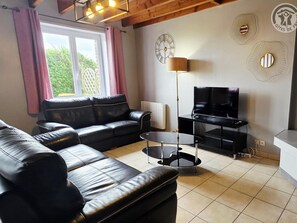 A living room with a black leather sofa, a coffee table, a TV on a stand, and decorative wall clocks. - Cozy house, private parking - 6 guests (Méteren)