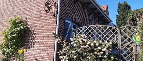 En byggnad av tegel med en grind av metall, ett staket av träribbor och en blommande buske.