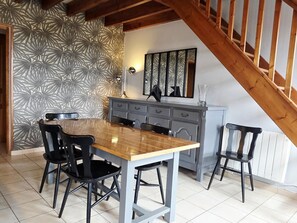 Un espace repas comprenant une table en bois, des chaises noires, un meuble gris et un escalier.