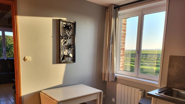 Een compacte keuken met een witte tafel, twee krukken, een radiator en een raam met uitzicht op groen.