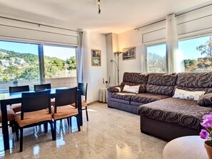 A living room with a sofa, a dining table with chairs, and a view of a hillside. - Cozy apartment near center - 5 guests (Tossa de Mar)