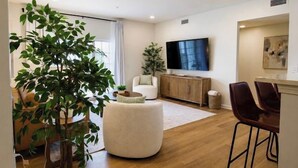 Un salon moderne avec un grand téléviseur, un pouf rond blanc, un canapé et un coin repas avec deux chaises.