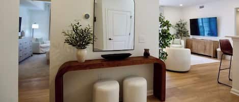 Una sala de estar moderna con un aparador de madera, dos otomanas blancas, un espejo grande y un televisor de pantalla plana.