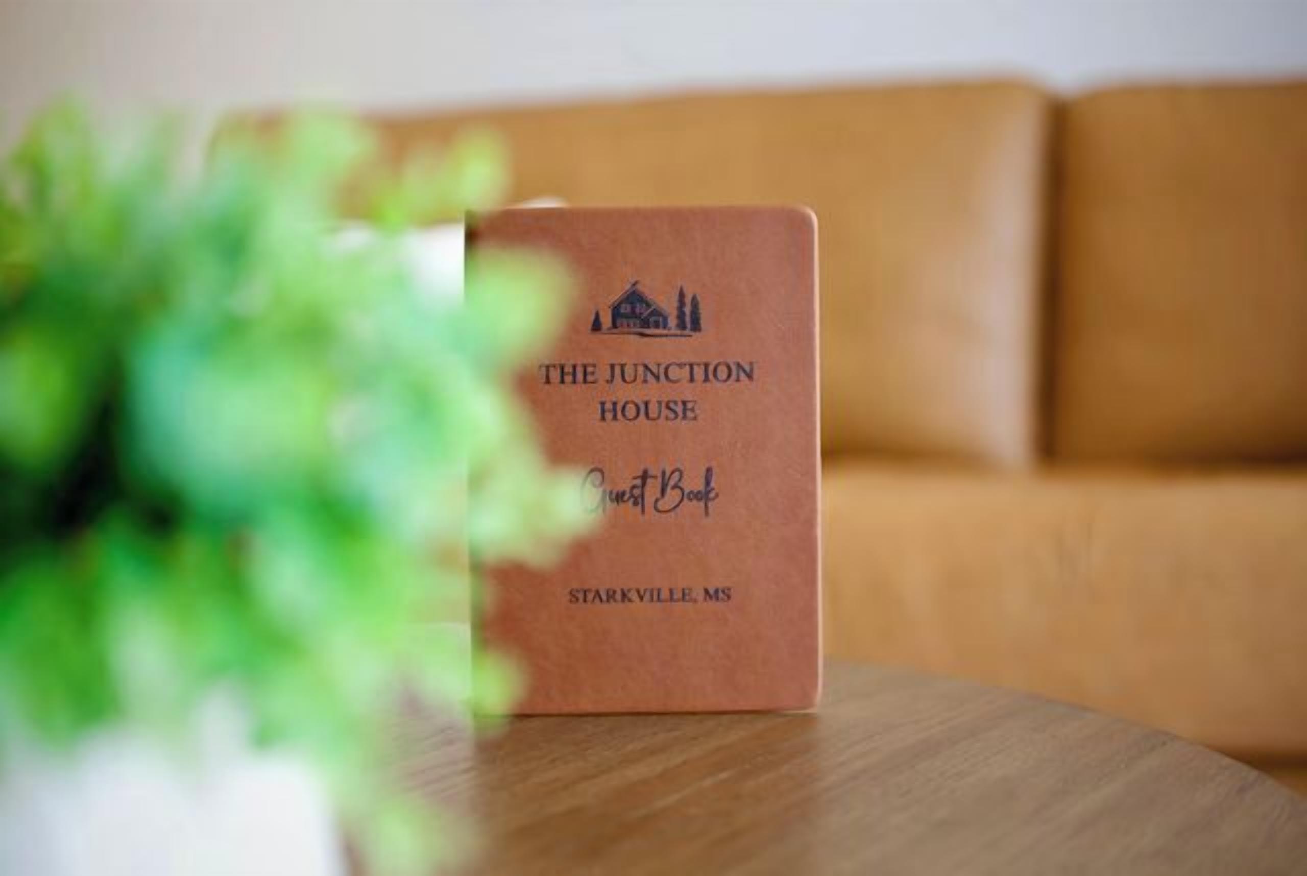 Ein braunes Buch mit dem Titel 'The Junction House' auf einem Holztisch, im Vordergrund verschwommen eine grüne Pflanze.