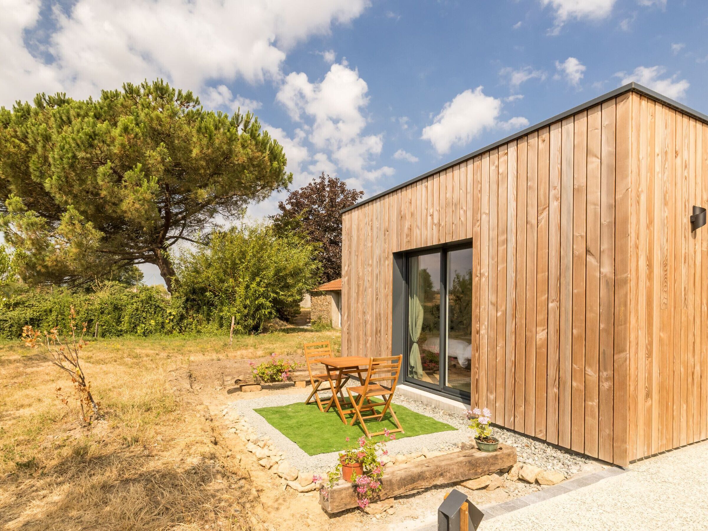 Ein modernes Holzgebäude mit Glastür, einer Terrasse mit Tisch und Stühlen und einem großen Baum im Hintergrund.