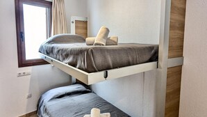 Una cama litera con colchón y almohadas, una ventana con cortinas y un estante fijado a la pared.