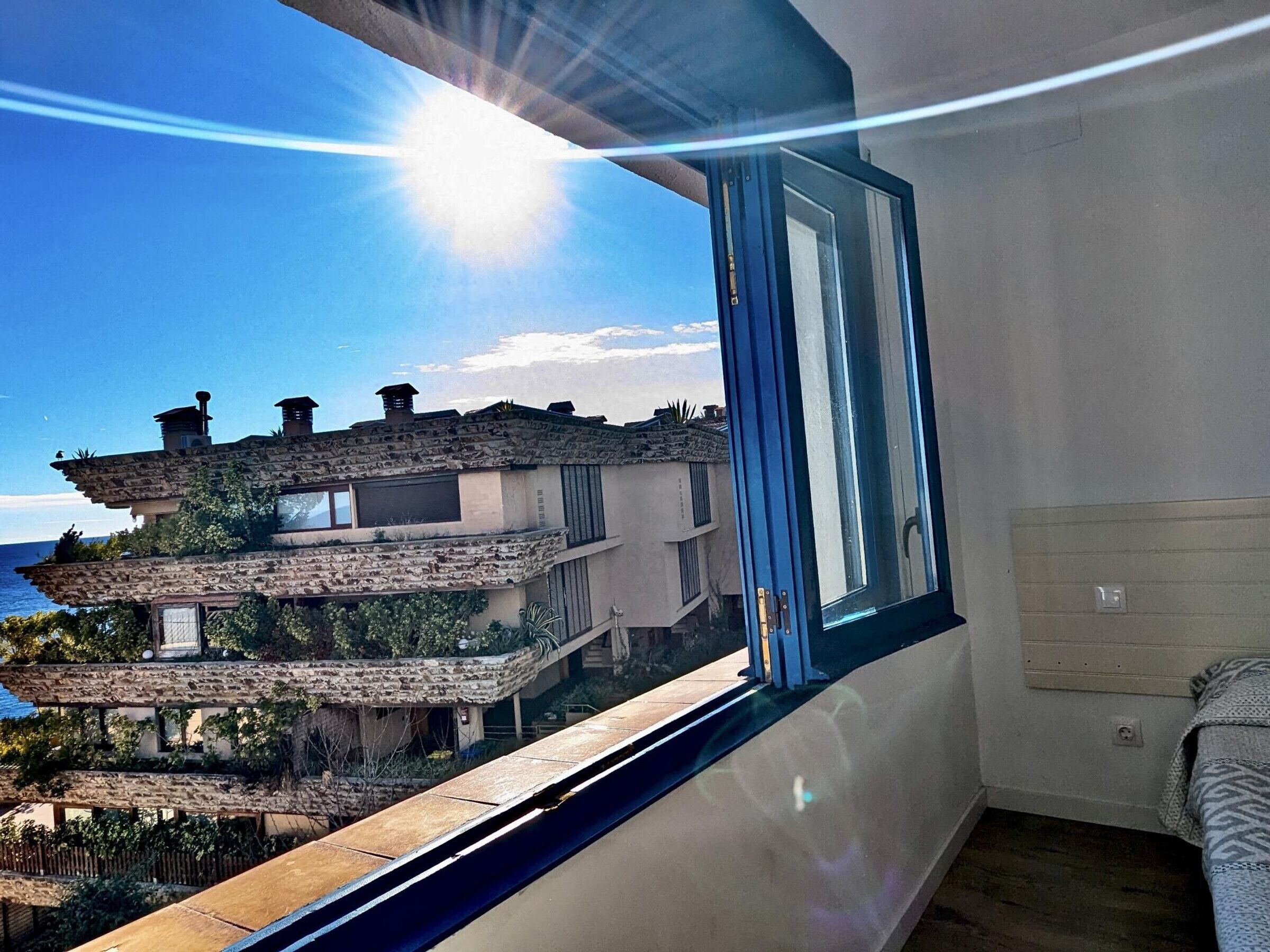 Ein Zimmer mit großem Fenster mit Blick auf ein Gebäude an der Küste, Holzboden und ein Bett mit einem gemusterten Überwurf.