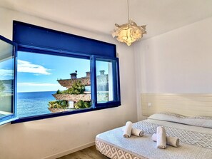 Ein Zimmer mit großem Fenster und Blick auf die Küste, einem Bett mit gemusterten Betttextilien und einer besonderen Lampenleuchte.