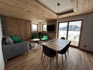 Ein modernes Interieur mit Holzbalkendecke, einem Esstisch mit Stühlen, einem Sofa mit grünen Kissen und Blick auf Berge durch ein großes Fenster.