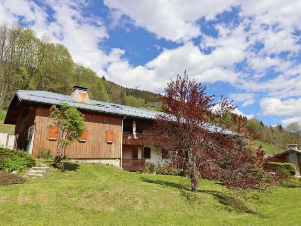 A wooden chalet with a sloped roof, a balcony, and a view of a forested hillside. - Apartment with parking - 4 guests (Les Gets)