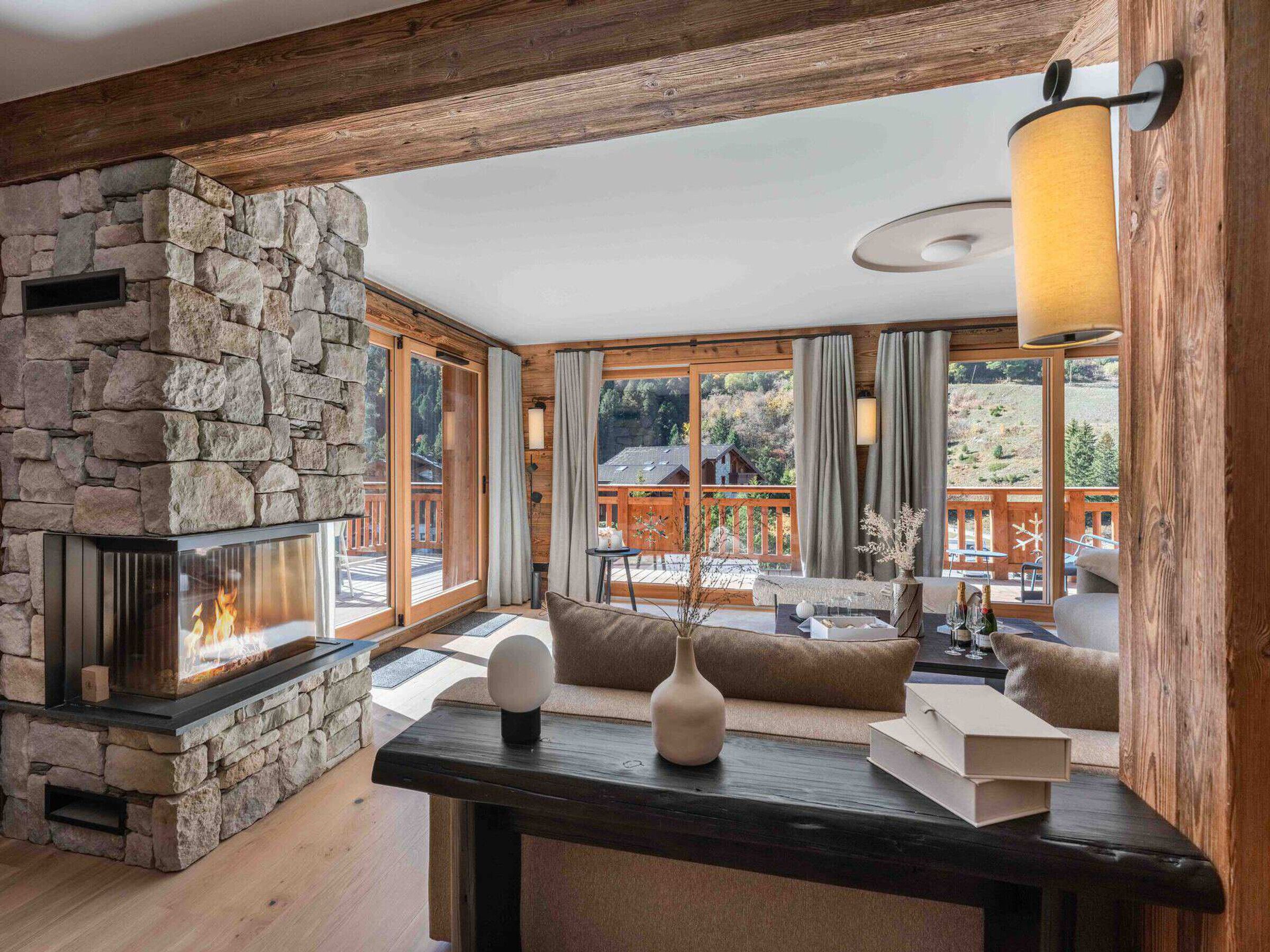 A cozy living room with a stone fireplace, wooden beams, large windows, and a view of a wooden deck.