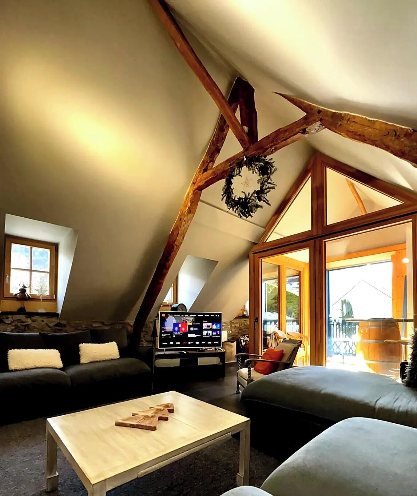 Un salon chaleureux de style loft, doté d’un plafond en pente, de poutres en bois, d’une cheminée et d’une grande fenêtre.