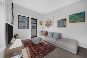 Un salon moderne avec un canapé, une table basse et un téléviseur. Les murs sont ornés de tableaux et il y a une fenêtre avec des stores.
