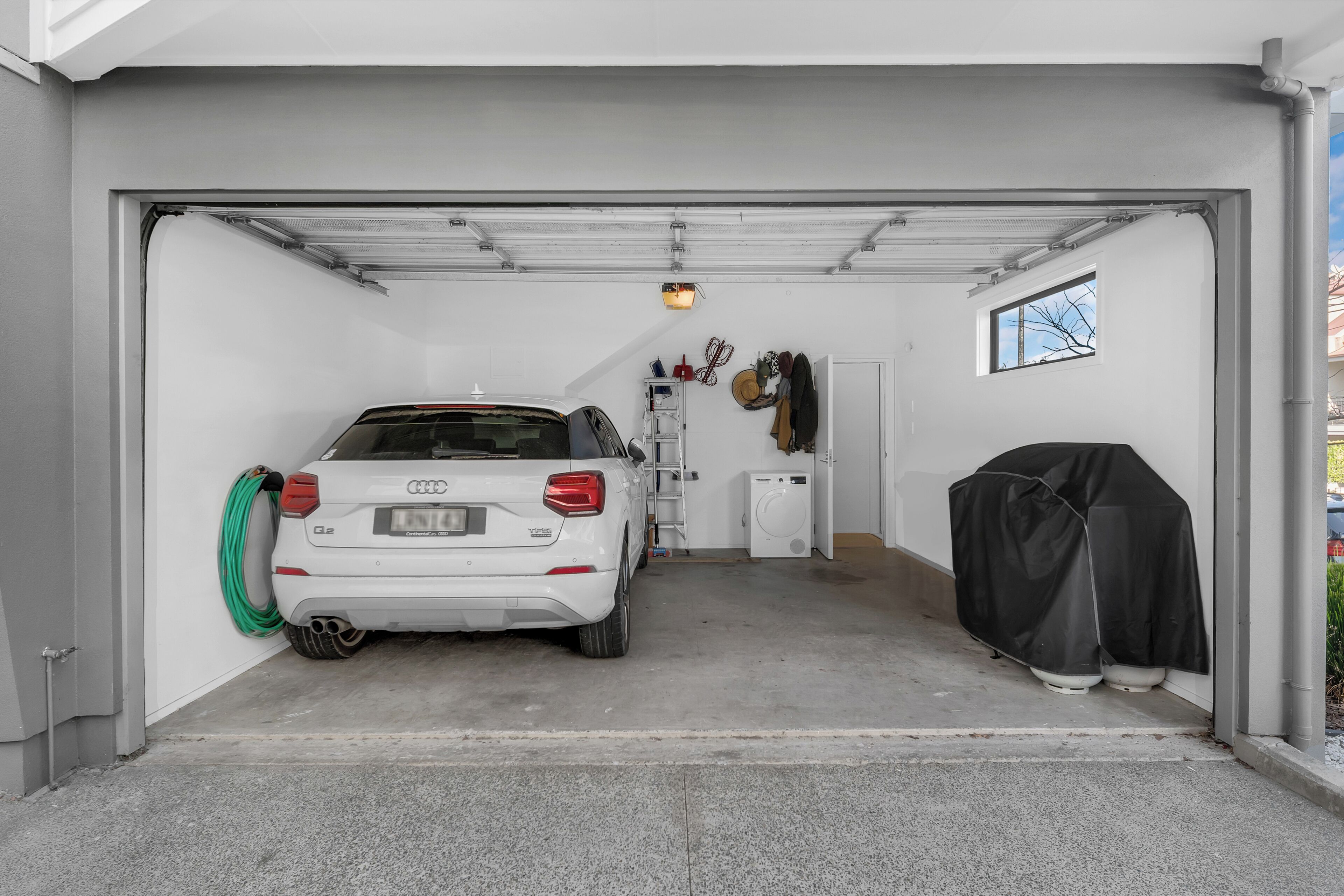 Une voiture blanche est garée à l’intérieur d’un garage, avec une machine à laver et un objet recouvert d’une housse sur la droite.