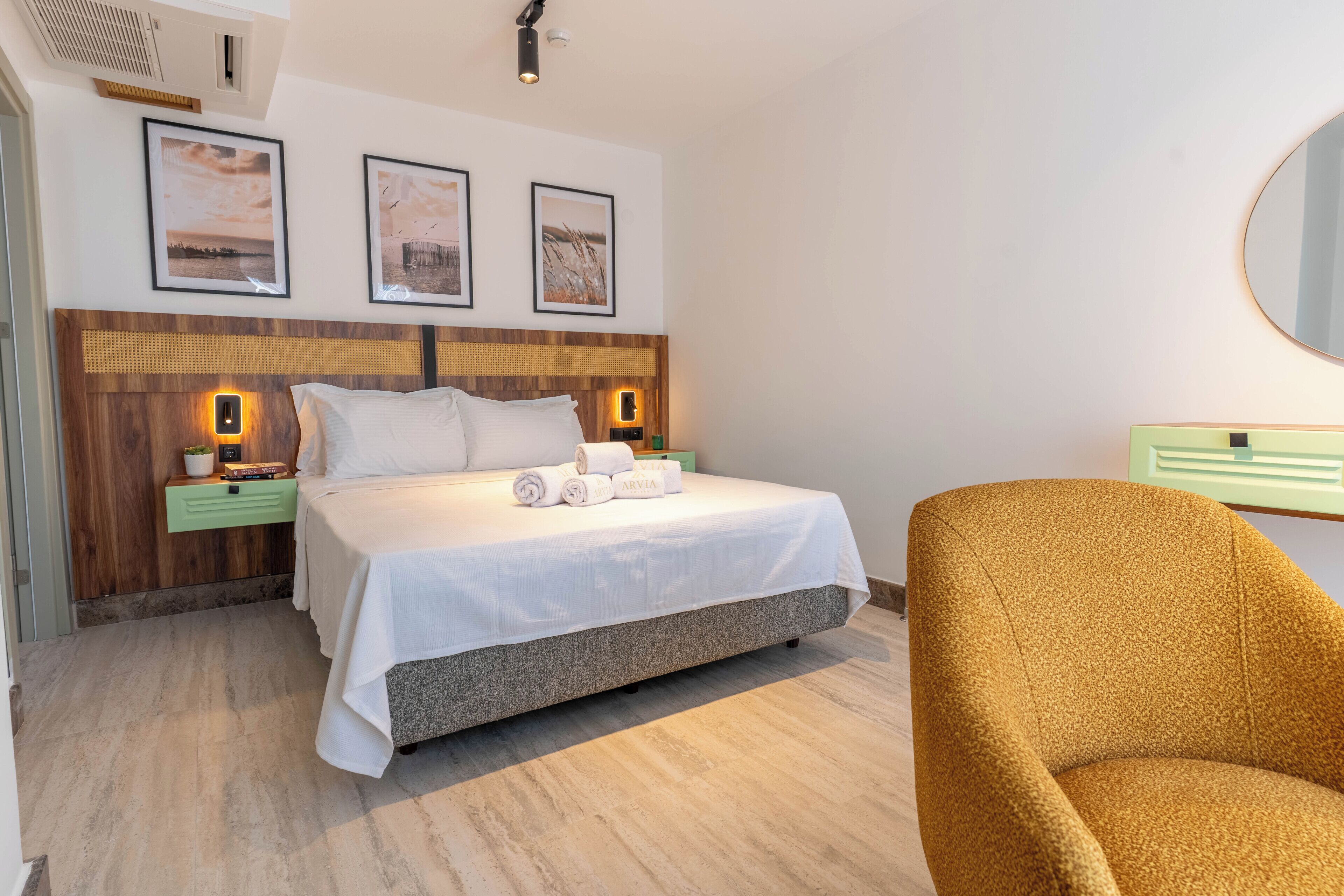 A hotel room with a bed, a nightstand, a green drawer, a round mirror, and a brown armchair.