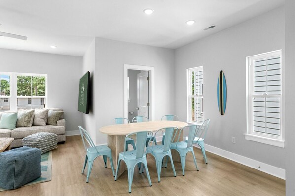 Uma sala de jantar com mesa de madeira e seis cadeiras, um sofá com almofadas, e vista para o exterior.