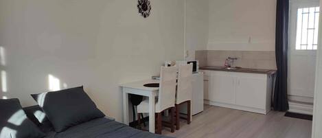 Ein kompakter Raum mit einem Bett, einem kleinen Esstisch mit Stühlen, einer Küchenzeile und einem Fenster mit Vorhängen.