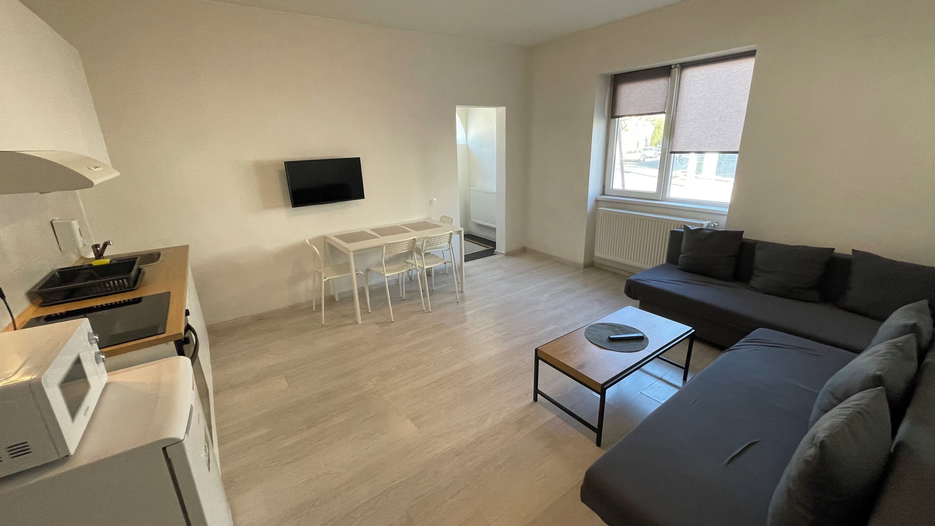 Ein modernes Wohnzimmer mit grauem Sofa, einem kleinen Couchtisch, einem an der Wand montierten Fernseher, einem Esstisch mit Stühlen und einem Fenster mit Jalousien.